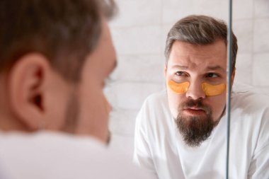 Portrait of man in white tshirt applying eye patches on his face at bathroom. Self care morning procedure. Beauty treatment concept