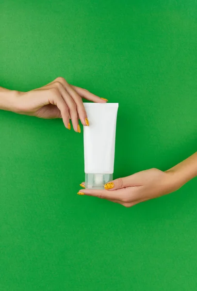 Female hands holding tube of cream lotion on green background. Cosmetic products blank package. Skin care routine.