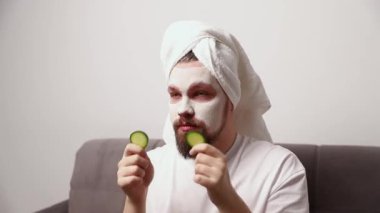 Beyaz tişörtlü komik bir adamın portresi yüzüne salatalık dilimleri sürüyor. Kişisel bakım sabah spa prosedürü. Güzellik tedavi konsepti