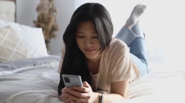 Yatağa uzanmış akıllı telefon kullanan sıradan giysili güzel Asyalı kadın. Oyun alışverişi çevrimiçi dinlendirici.