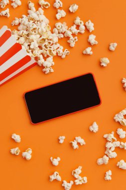 Turuncu arka planda akıllı telefon ve bir kutu patlamış mısır. Sinema, sinema ve eğlence konsepti.