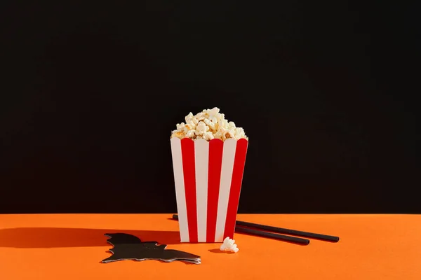 Striped box with popcorn, bat napkin and straws on an orange black background. Halloween movie night