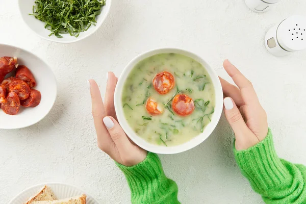 Womens hands holding bowls of Portuguese style soup Caldo Verde with bread, cabbage, and chorizo sausage on white background. Traditional Portuguese food.