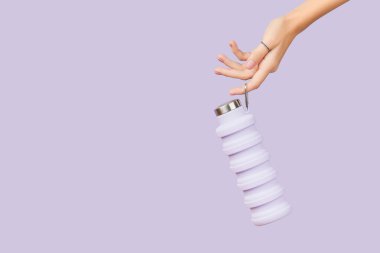 Kadınların elleri, mor arka planda, yeniden kullanılabilir, katlanabilir leylak su şişesini tutuyor. Sıfır atık, sürdürülebilir yaşam tarzı