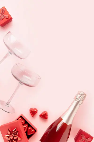 A bottle of sparkling wine with two glasses and heart shaped chocolate candies in red gist box on pink background with copy space. Valentines Day concept. Flat lay
