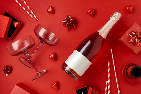 A bottle of sparkling wine with two glasses, red gift boxes with ribbons and heart shaped chocolate candies on red background. Valentines Day concept. Flat lay