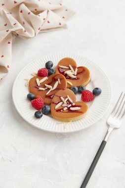 İçinde ahududu, yaban mersini, badem gevreği ve kırmızı böğürtlen sosu olan bir yığın kalp şeklinde krep ve gri arka planda beyaz tabakta çatal ve kalp baskılı doku. Sevgililer Günü konsepti 