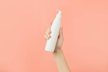 A womans hand holding a white cosmetic bottle on a peach background. Care products beauty treatment concept