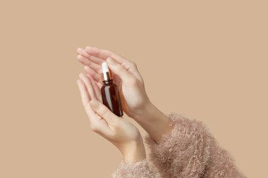 A womans hands holding a brown bottle of serum on a beige background. Body care beauty treatment concept