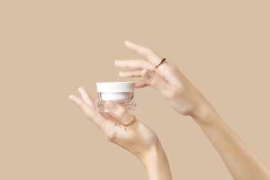 A womans hands holding a white cosmetic jar on a beige background. Care products beauty treatment concept