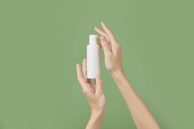 A womans hands holding a white cosmetic bottle on a green background. Morning skincare routine