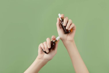 A womans hands holding a brown bottles of serum on a green background. Morning skincare routine