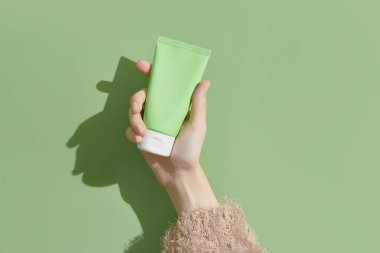 A womans hand in a sweater holds a tube of cream on a green background. Self care beauty treatment concept