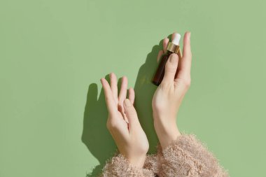 A womans hands holding a brown bottle of serum on a green background. Morning skincare routine