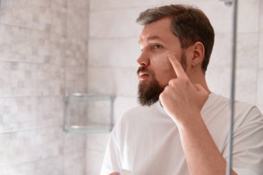 Beyaz tişörtlü bir adamın portresi banyoda yüzüne krem sürüyordu. Kişisel bakım sabah prosedürü. Güzellik tedavi konsepti