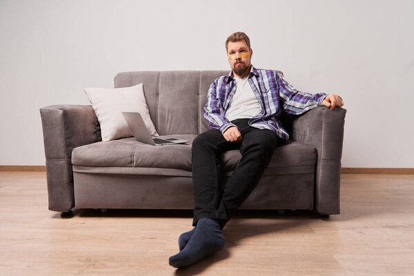 Человек в фиолетовой рубашке с повязками на глазу сидит на диване с ноутбуком. Косметические процедуры spa wellbeing концепция. Самообслуживание