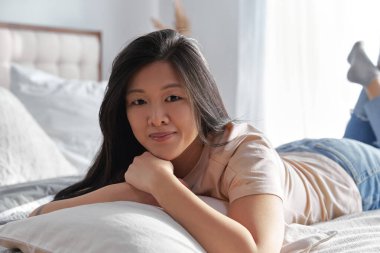Mutlu Asyalı kadın gündelik giysiler içinde evde yatakta yatıyor. Rahatlama konseptinin tadını çıkarın.