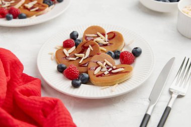 Ahududu, yaban mersini, badem gevreği, beyaz tabakta kırmızı böğürtlen sosu, çatal, bıçak ve gri arka planda kırmızı peçete olan bir yığın kalp şeklinde krep. Sevgililer Günü konsepti 