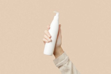 A womans hand holding a white cosmetic bottle on a beige background. Care products beauty treatment concept