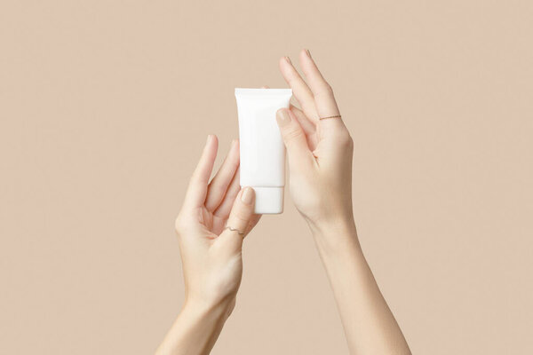 A womans hands holding a white cosmetic tube on a beige background. Care products beauty treatment concept