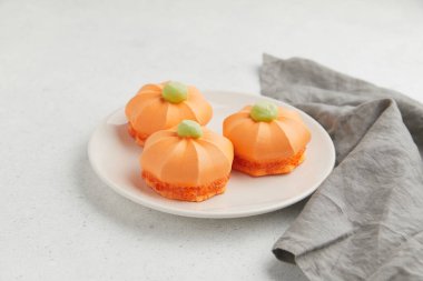 Üç turuncu balkabağı şekilli yeşil kapaklı tatlılar, yakınlarda gri bir bez parçasıyla beyaz bir tabağa yerleştirilmiş.