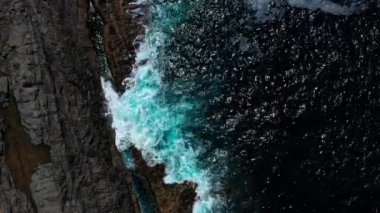 Okyanus dalgalarının kayalara çarpmasının havadan görüntüsü. Yukarıdan aşağıya dron görüntüsü okyanus arka planını dalgalandırıyor. Ada kayalıklarında su sıçrayan ve köpüren dalgalar. Gökyüzünden güzel bir deniz manzarası