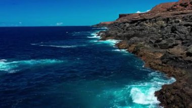 Derin mavi suyun üzerinde uçan bir dron. Güçlü dalgalar Fuerteventura adasındaki siyah volkanik kayalara çarpıyor ve köpürüyor. Seyahat ve tatil kavramı.