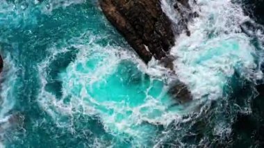 Okyanus dalgalarının kayalara çarpmasının havadan görüntüsü. Yukarıdan aşağıya dron görüntüsü okyanus arka planını dalgalandırıyor. Ada kayalıklarında su sıçrayan ve köpüren dalgalar. Gökyüzünden güzel bir deniz manzarası.