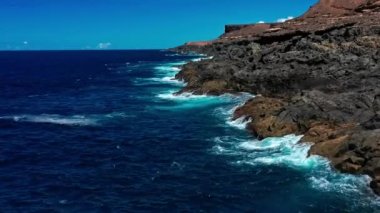 Derin mavi suyun üzerinde uçan bir dron. Güçlü dalgalar Fuerteventura adasındaki siyah volkanik kayalara çarpıyor ve köpürüyor. Seyahat ve tatil kavramı.