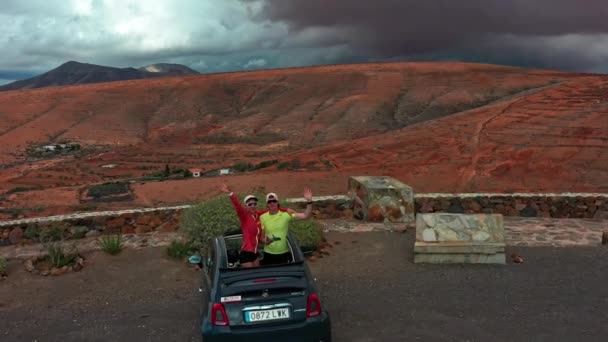 来自高加索的男女游客从车上挥手而下 无人驾驶飞机飞越了他们的上空 展现了一座富尔特文图拉火山岛上美丽的山地风景 背景是多彩多姿的云天 — 图库视频影像