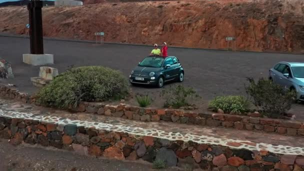 白人男女游客从车上挥手而下 无人驾驶飞机从他们身边飞走 展现了一座富尔特文图拉火山岛上美丽的山地风景 — 图库视频影像