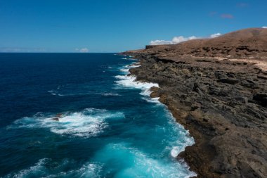 Derin mavi suyu olan güzel bir manzara. Güçlü dalgalar Fuerteventura adası Aguas Verdes sahilindeki siyah volkanik kayalara çarpar. Seçici odak, bulanık arkaplan.