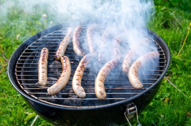 Sizzling Sausages on Outdoor Barbecue Grill. Smoky Bratwurst Cooking Over Charcoal in Sunny Backyard clipart