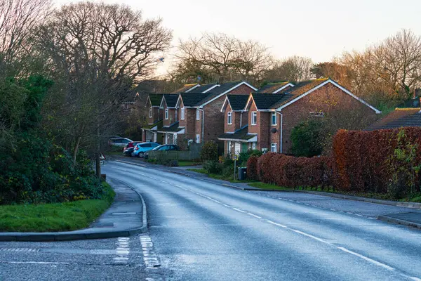 Takeley, İngiltere - 7 Aralık 2024: Tuğladan evler ve kış ağaçları olan modern yerleşim caddesi, Takeley, Essex. Park etmiş arabalarla kavisli banliyö yolu ve alacakaranlıkta tipik İngiliz mimarisi.
