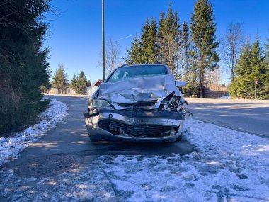 Kış sokağına park edilmiş büyük bir çarpışma hasarı olan gümüş renkli bir araba. Çarpışan araç buruşuk kapüşonlu, ezilmiş tampon ve karlı yol kenarında kırık ızgara gösteriyor.