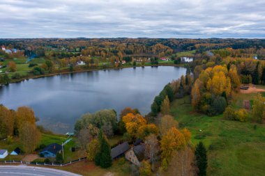 Estonya kırsalında canlı sonbahar yapraklarıyla çevrili Rouge Gölü 'nün havadan görünüşü. Kırsal köy, kıyı boyunca dağılmış evler bulutlu gökyüzünün altında Estonya 'nın güzelliğini gözler önüne seriyor.