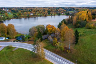 Estonya kırsalında canlı sonbahar yapraklarıyla çevrili Rouge Gölü 'nün havadan görünüşü. Kırsal köy, kıyı boyunca dağılmış evler bulutlu gökyüzünün altında Estonya 'nın güzelliğini gözler önüne seriyor.