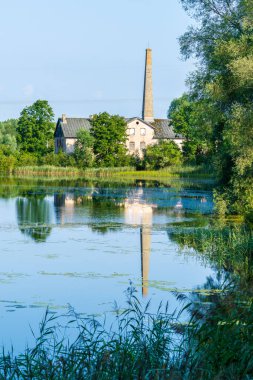 Sakin nehir suyuna yansıyan uzun bacalı tarihi tuğla fabrikası. Eski endüstriyel değirmen binası yemyeşil ağaçlar ve sazlıklarla çevrili.