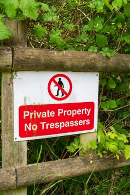 Kırmızı ve beyaz yasaklama işareti, kırsal bir ahşap çite bağlı özel araziye izinsiz girilmemesi konusunda uyarıda bulunuyor. Yabani bitki örtüsüyle çevrili mülk sınır uyarısı