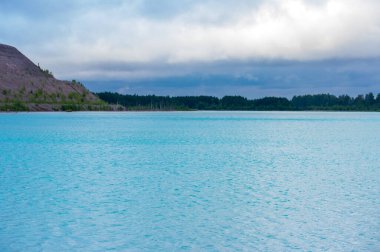 Orman ve tepelerle çevrili güzel mavi su gölü dramatik bir gökyüzünün altında. Turkuaz su ve ormanlık alanla sakin doğal manzara