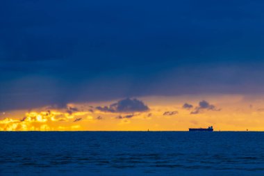 Kargo gemisi silueti okyanus ufkunda dramatik günbatımında altın bulutlar ve derin mavi gökyüzü ile. Renkli alacakaranlıkta canlı ışık gösterisiyle durgun suda seyreden bir gemiyle güzel bir deniz manzarası.