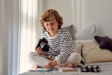 Dijital fotoğraf makineli okul çocuğu yatak odasında bacak bacak üstüne atmış otururken not alıyor.