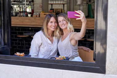 Gündelik giysiler içinde, pencerenin yanında otururken akıllı telefondan selfie çeken ve modern kafede lezzetli yemekler yiyen pozitif genç kadınlar.