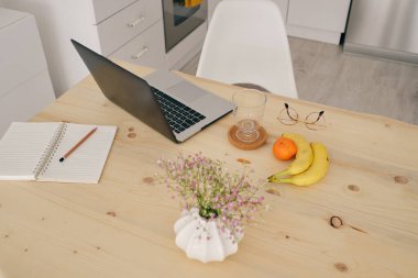 Yüksek açılı netbook, ahşap masanın üzerine vazoda çiçek buketi ve evdeki mutfakta kalemle birlikte not defterinin yanında meyveler.