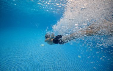 Şortlu, maske takan, temiz havuzda yüzen, mavi kartonla kaplı gömleksiz bir çocuk.