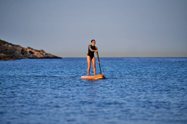 Mayo giymiş ince kadın sörfçülerin bütün vücudu SUP tahtasının üzerinde duruyor ve gün batımında dalgalı deniz suyunda kürek çekiyor ve gözlerini kaçırıyor.