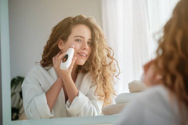 Sabahları aynaya bakarken modern elektrik süpürgesiyle bornozlu kadın cilt bakımı yapıyor.