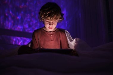 Gece vakti karanlık yatak odasında yatağında yatarken gece giysisi sörf tabletine odaklanmış bir çocuk.