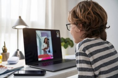 Hoş bir çocuğun, sınıf arkadaşıyla bilgisayar aracılığıyla telefon görüşmesi yaparken aydınlatma odasındaki akıllı telefonlu masada oturması yan görüntüsü.