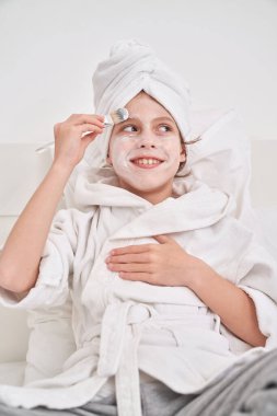 Yüzüne nemlendirici maske sürerken yatağın üzerine oturup yastıklara yaslanan bornoz ve havlu giymiş pozitif bir çocuk.